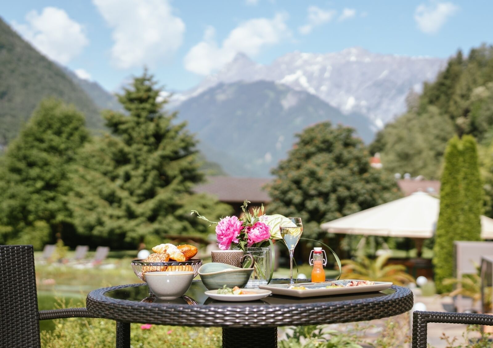 Ein Tisch gedeckt mit einer Schale Gebäck, einem Glas Weißwein und einer Flasche Orangensaft, mit Blick auf Berge im Hintergrund.