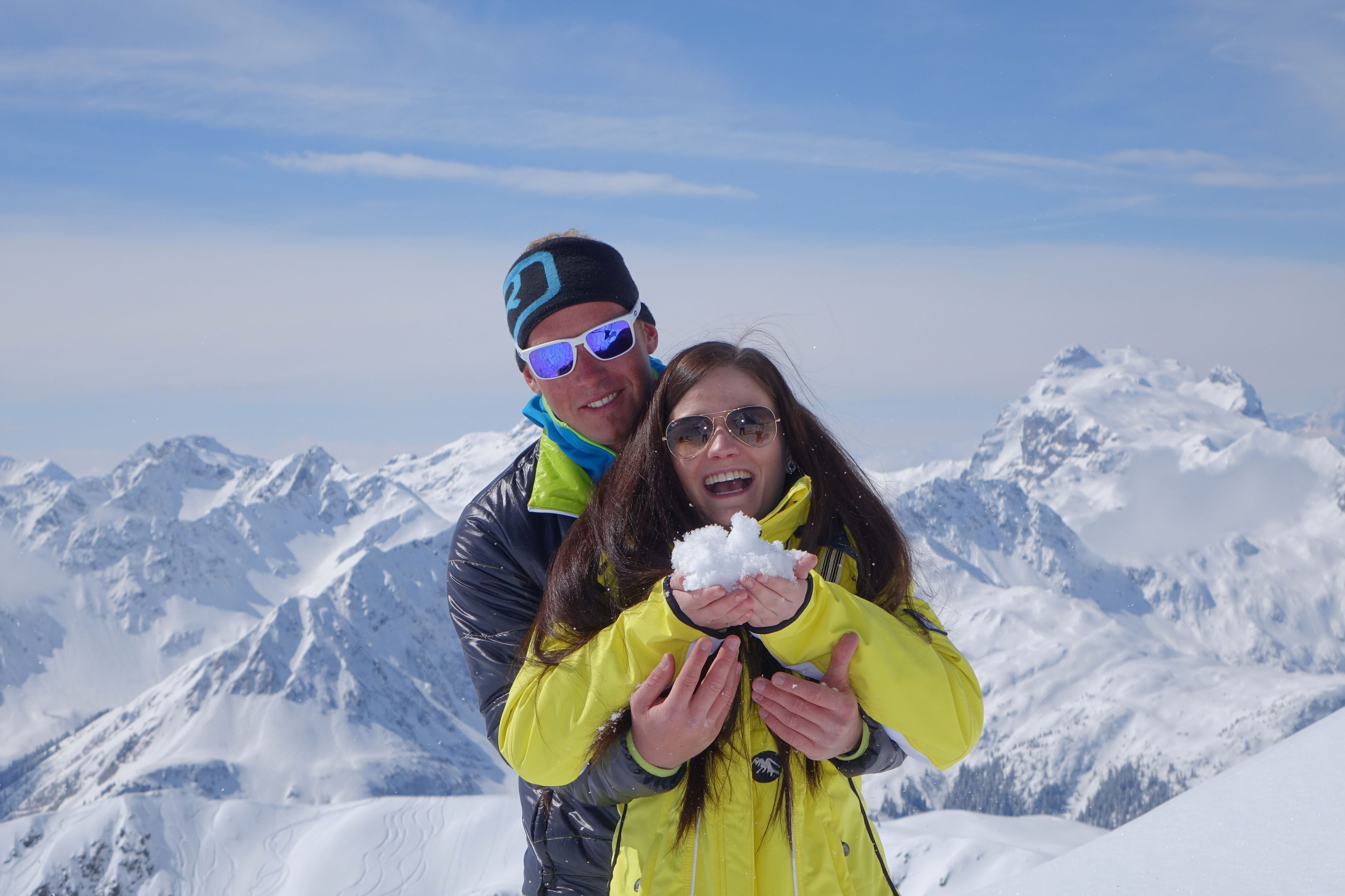 A couple standing on a snowy mountain with snow-covered peaks in the background.