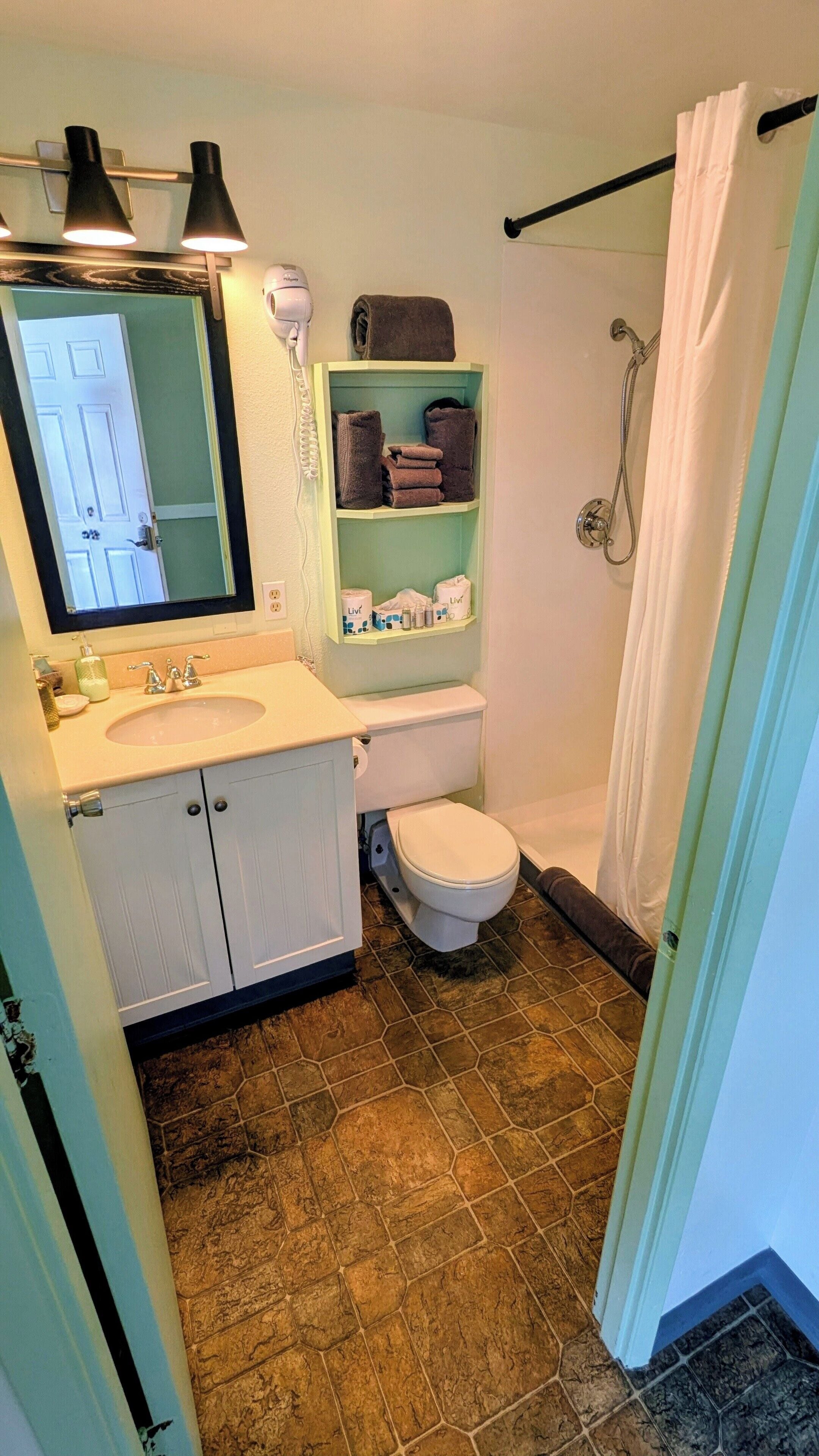 Une salle de bain équipée d’un WC, d’un lavabo, d’un miroir, d’une étagère avec des serviettes et d’un rideau de douche.