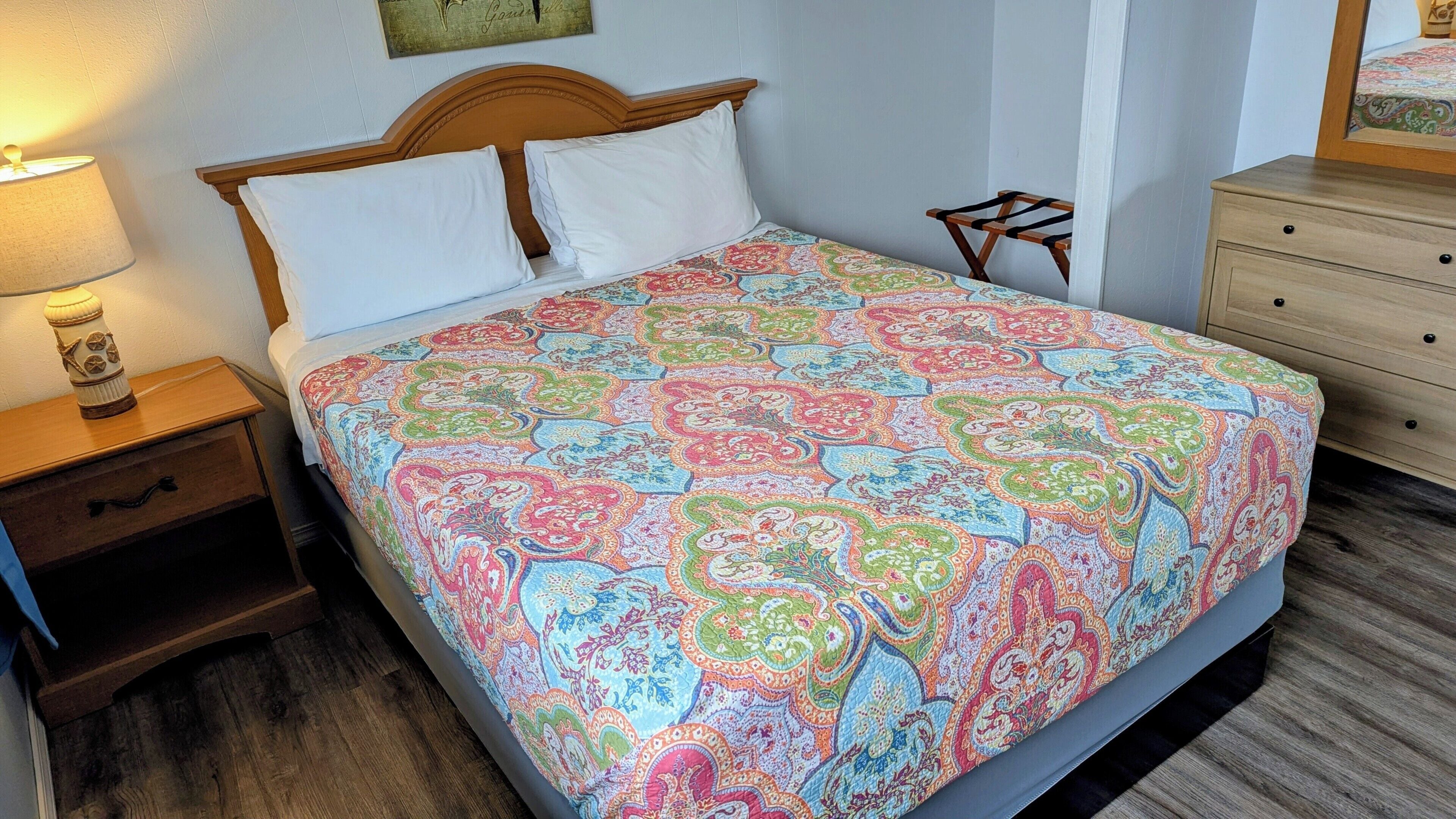 A neatly made bed with a patterned bedspread, a bedside lamp, a small wooden nightstand, and a dresser with drawers.