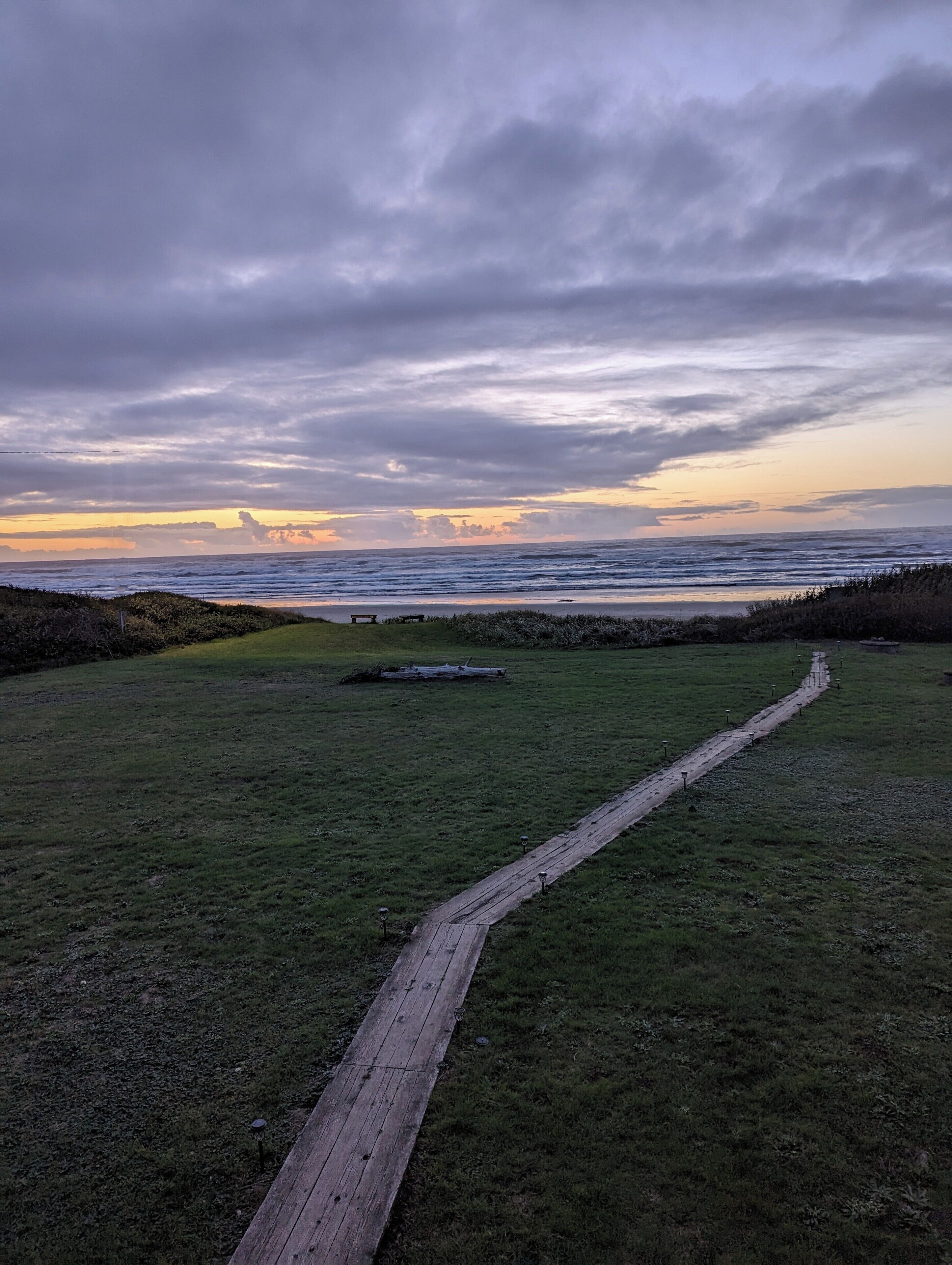 A grassy area with a wooden boardwalk leading to a bench, overlooking a body of water under a cloudy sky.