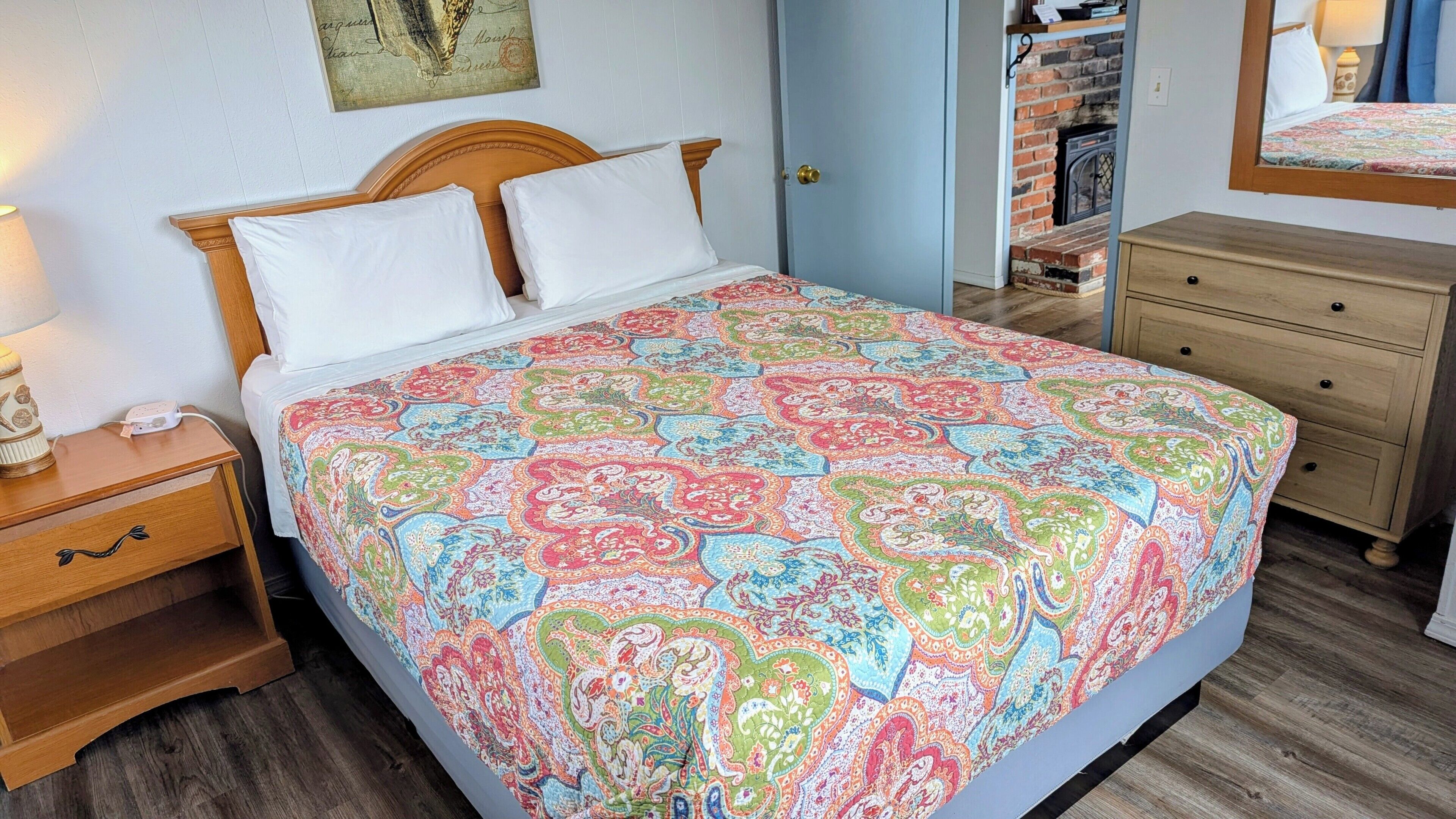A bed with a colorful patterned bedspread, a wooden nightstand with a lamp, a dresser, and a brick fireplace.