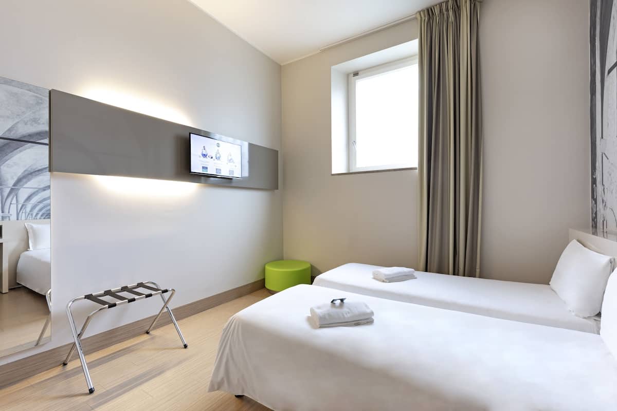 Ein Hotelzimmer mit zwei Betten, einem Wand-Fernseher, einem grünen Hocker und einem Fenster mit Vorhängen.