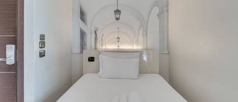A narrow hotel room with a bed, a nightstand, a mirror, and a window with a view of the interior.