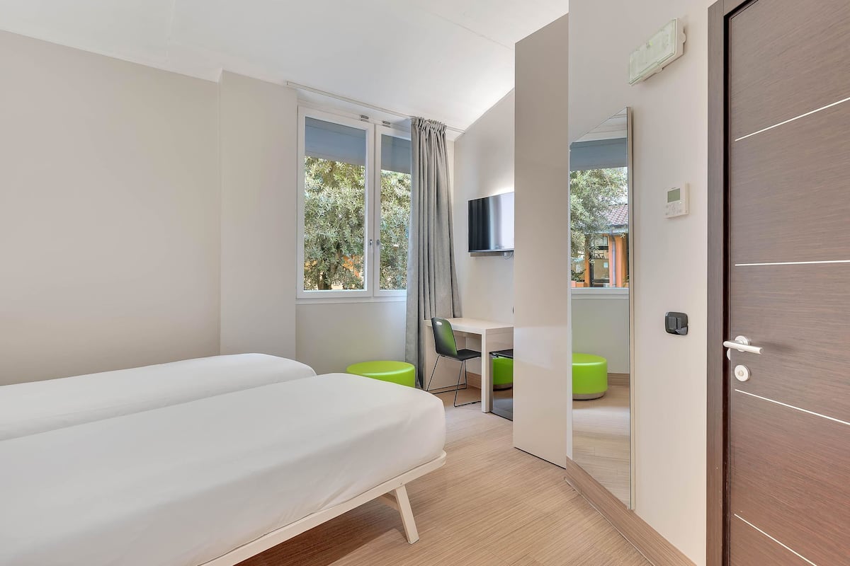 Ein modernes Hotelzimmer mit einem Bett, einem Schreibtisch, einem Stuhl und einem grünen Hocker.