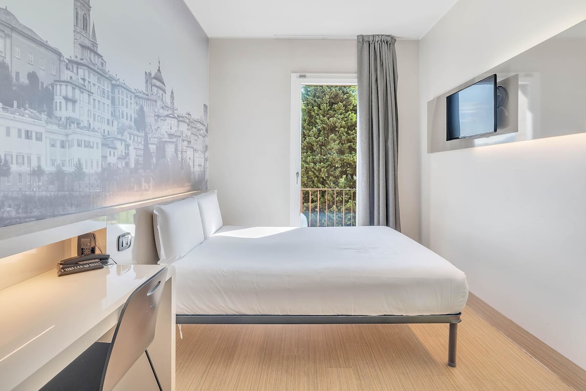 Ein Hotelzimmer mit Bett, Schreibtisch mit Telefon, Wandfernseher und Fenster mit Blick auf Bäume.