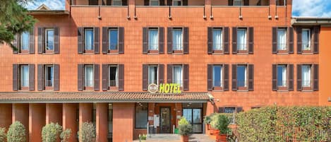 Un hotel de varios pisos con fachada roja, numerosas ventanas con contraventanas y una entrada central.