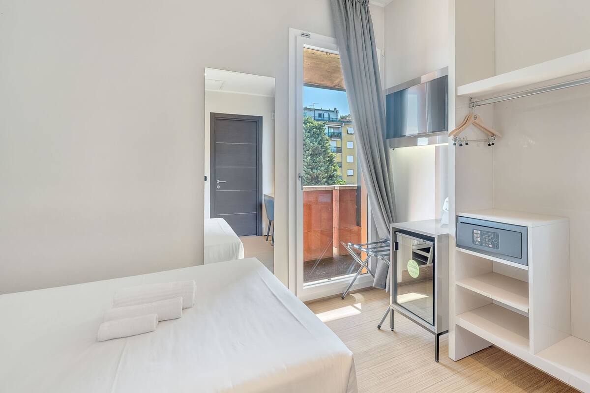 Ein modernes Hotelzimmer mit Bett, Schreibtisch mit Stuhl, Kleiderschrank und einem Balkon mit Blick auf Gebäude und Bäume.