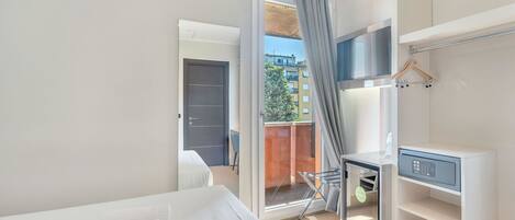 Una habitación de hotel moderna con cama, escritorio y silla, armario y balcón con vistas a edificios y árboles.