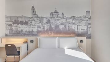 Camera d'albergo con un letto, una scrivania e una sedia. Dietro al letto si trova un grande murale che raffigura un panorama cittadino.