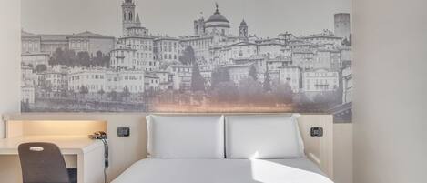 A hotel room with a bed, a desk, and a chair. There is a large cityscape mural on the wall behind the bed.