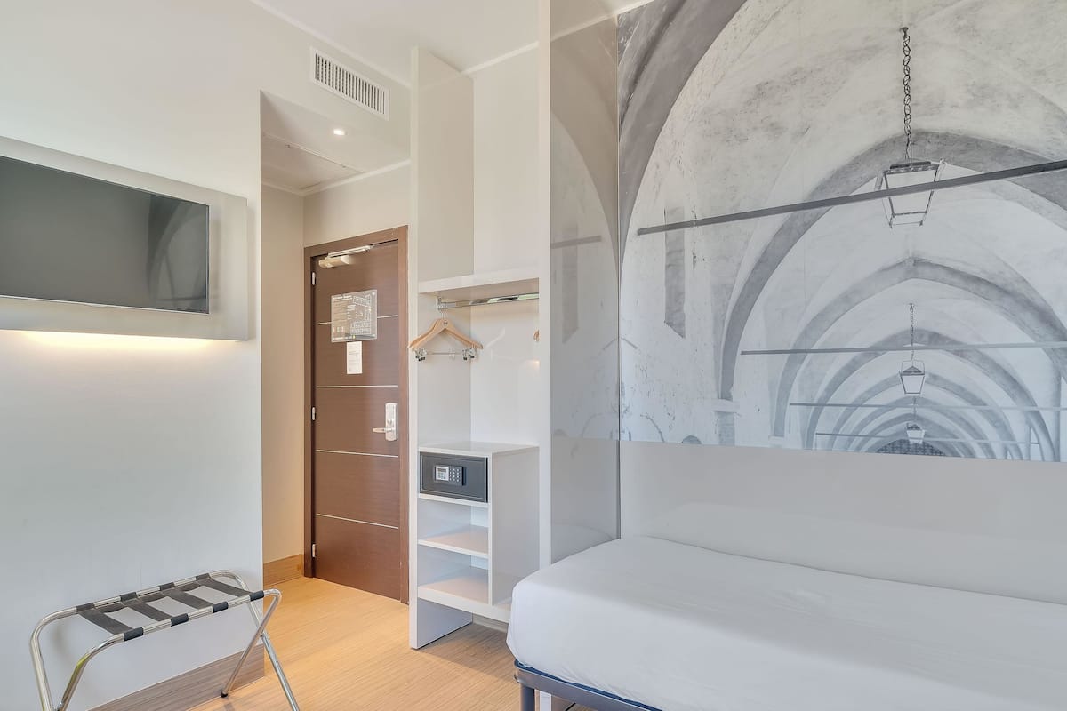 Ein modernes Hotelzimmer mit einem Bett, einem an der Wand montierten Fernseher, einem Kleiderständer und einem Safe.