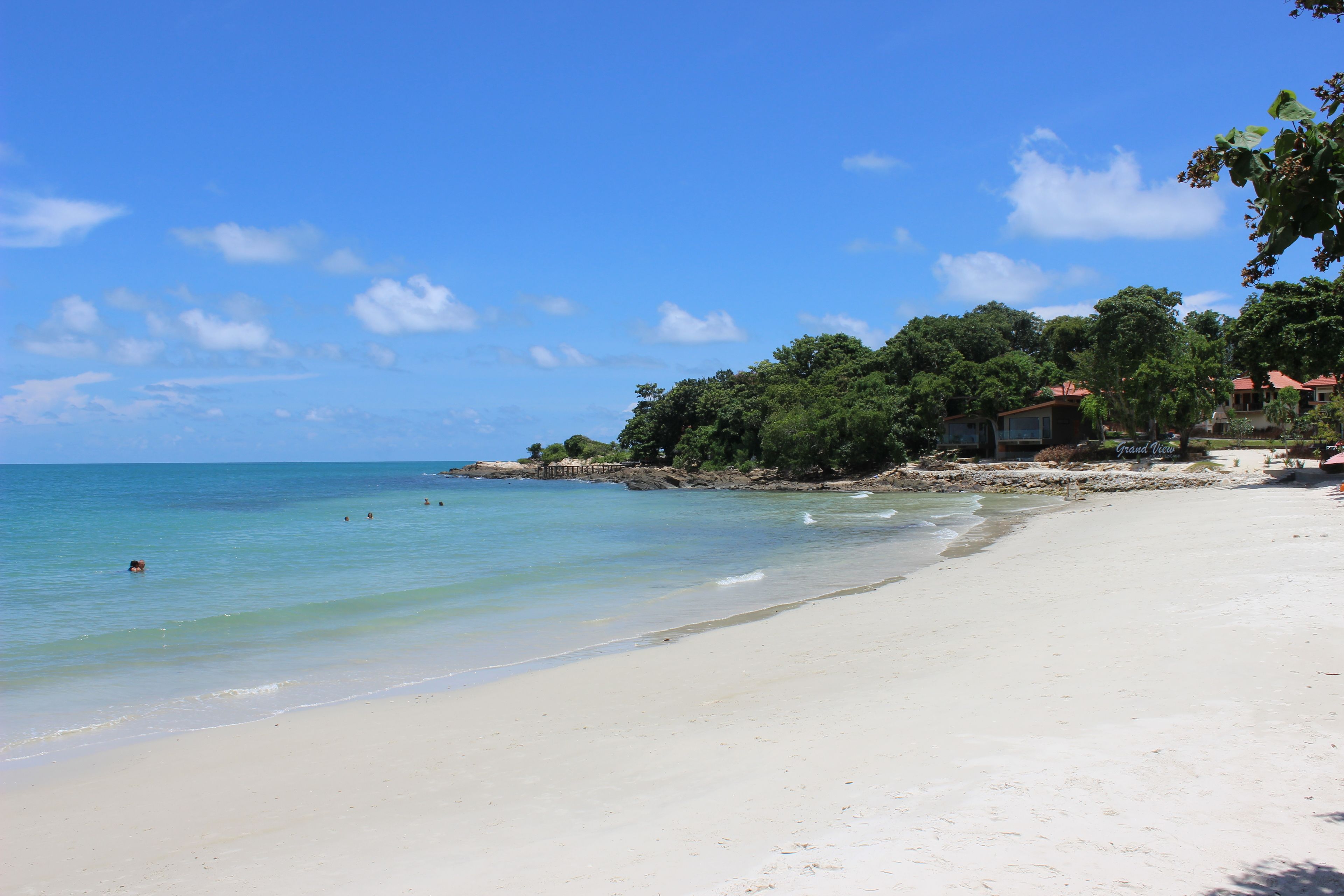 A beach with clear blue water, white sand, and a few people swimming. There are trees and buildings in the background.