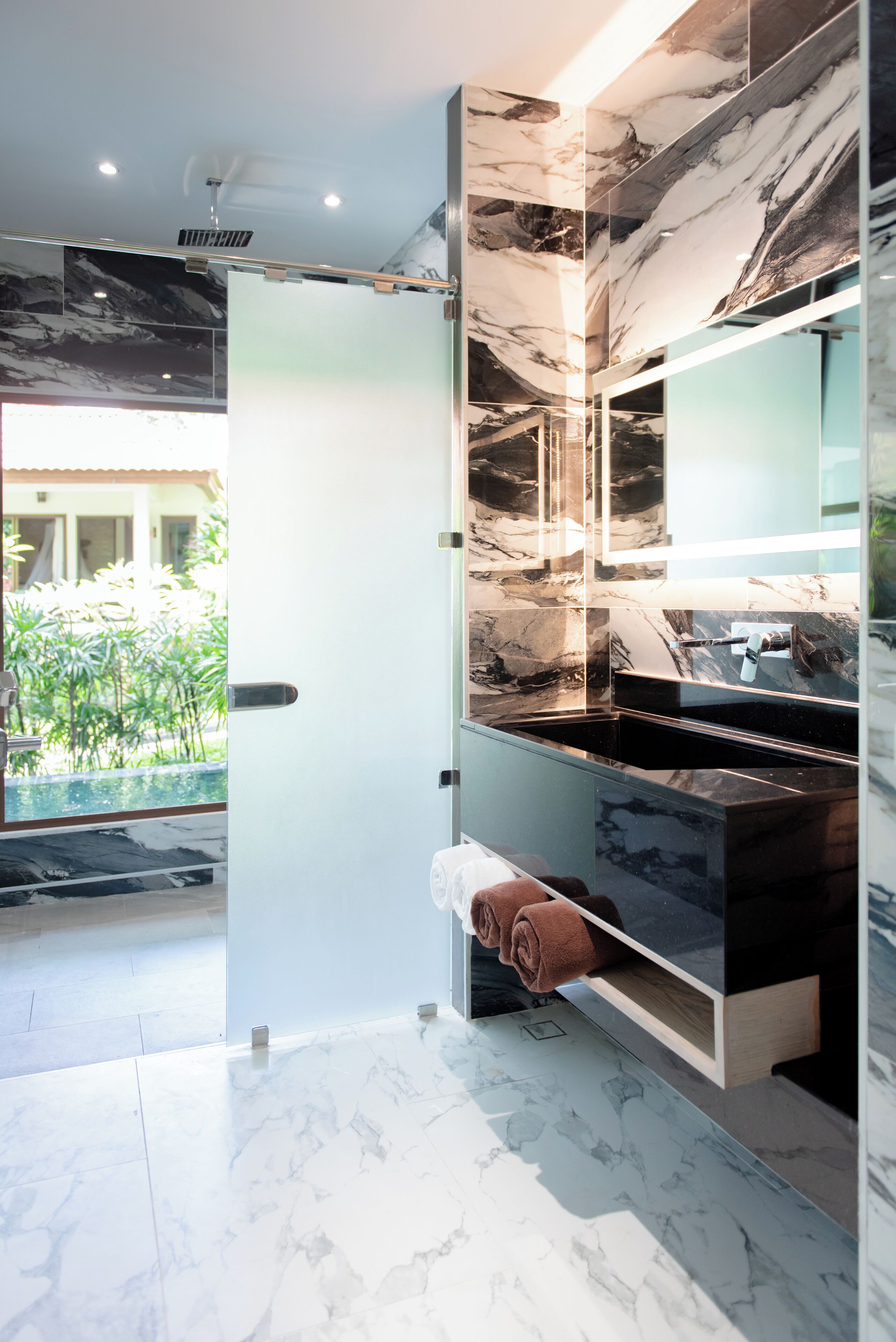 Un baño moderno con tocador de mármol, cabina de ducha de vidrio y vista a un jardín.