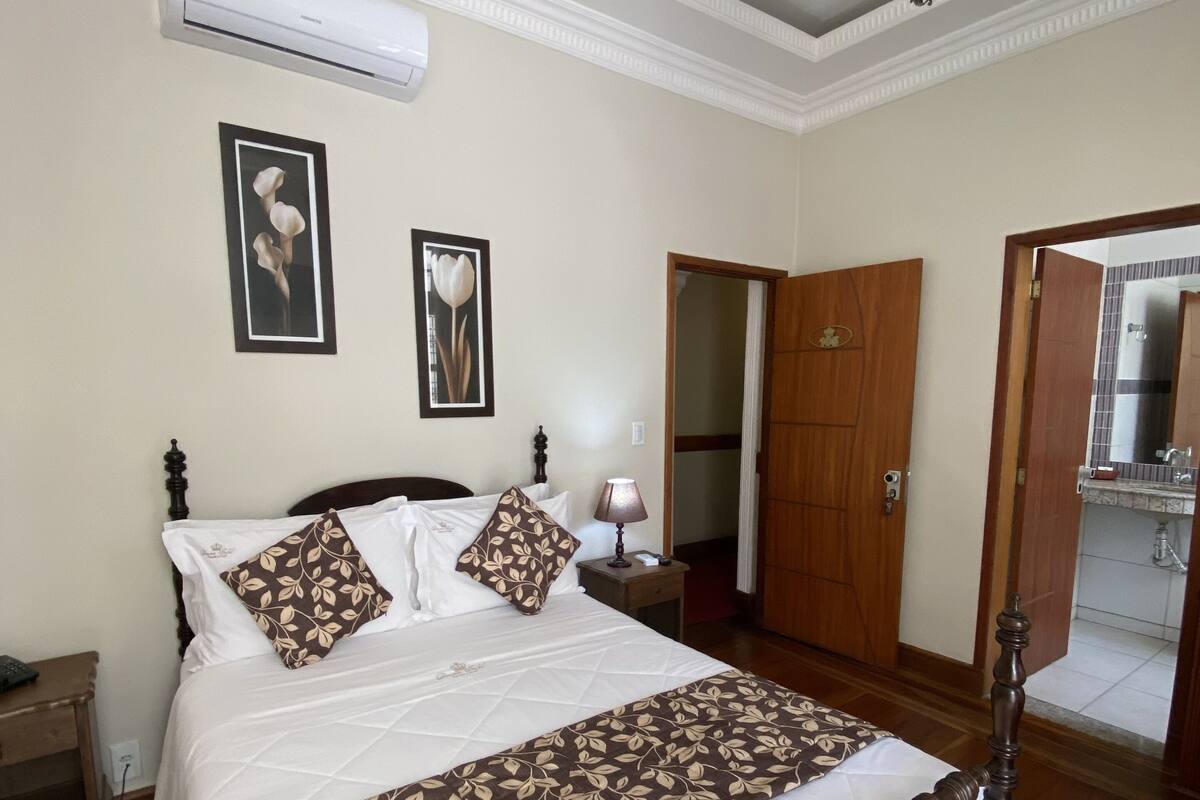 Quarto com cama de dossel, lustre, aparelho de ar condicionado e duas molduras na parede.