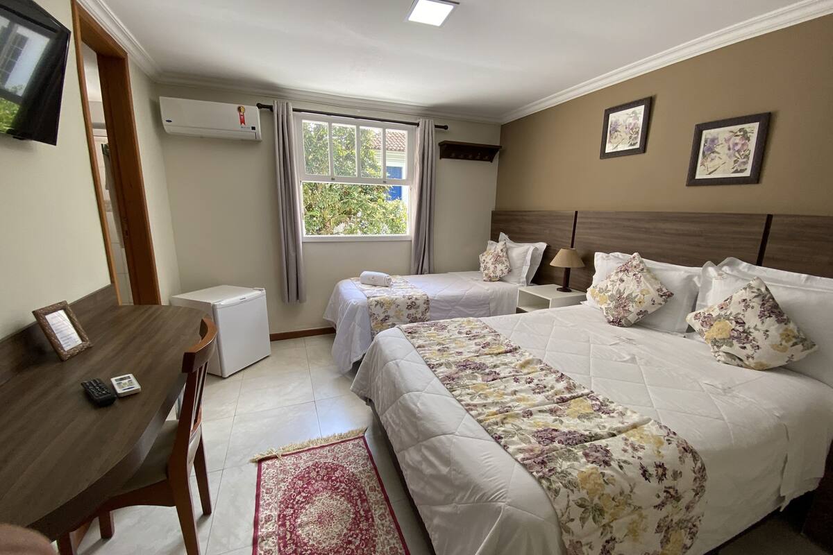 Quarto de hotel com duas camas, uma escrivaninha, uma cadeira, um refrigerador pequeno e uma janela com cortinas.