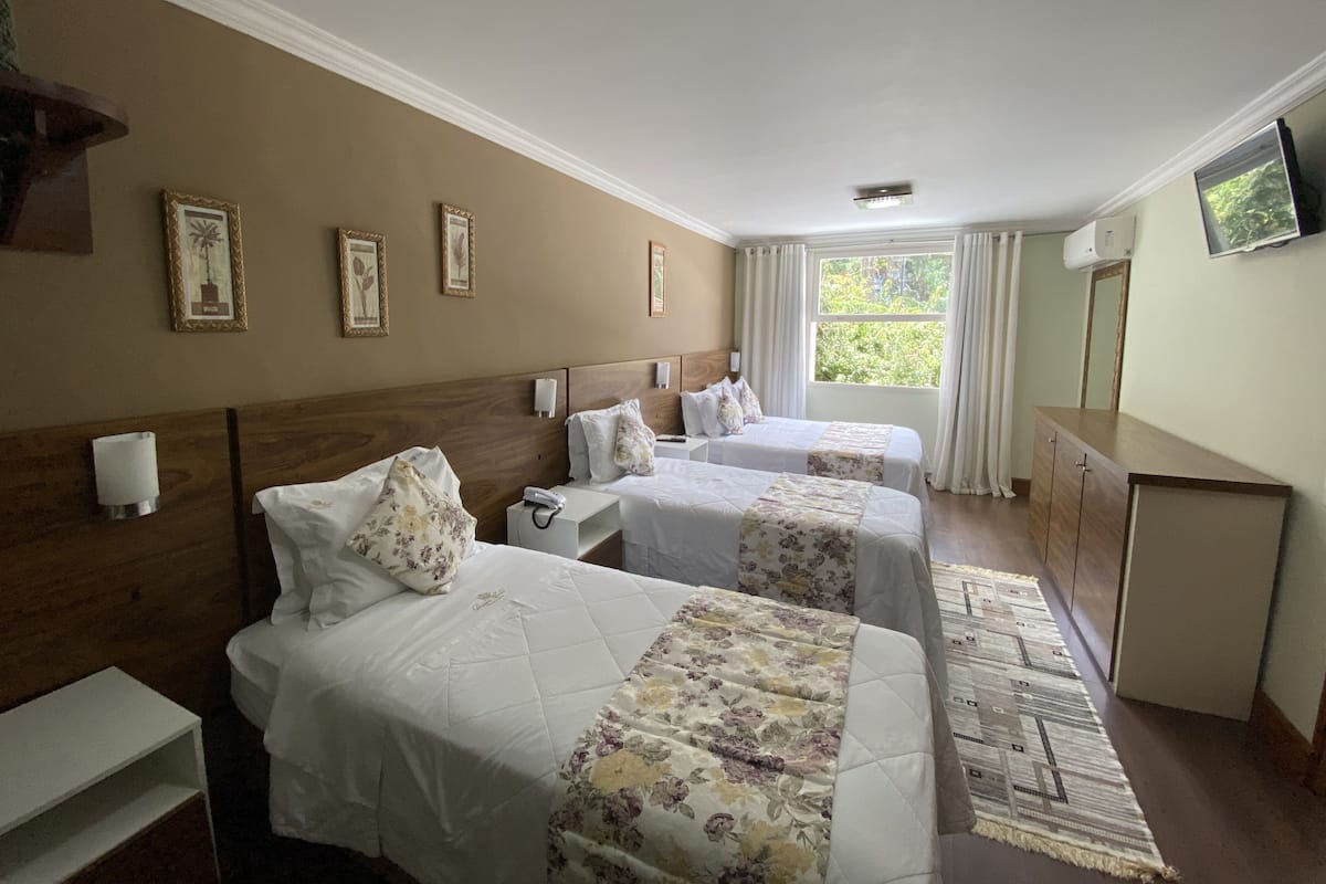 Quarto de hotel com duas camas, uma cabeceira de madeira, mesa de cabeceira com abajur, espelho, televisão na parede e janela com cortinas.