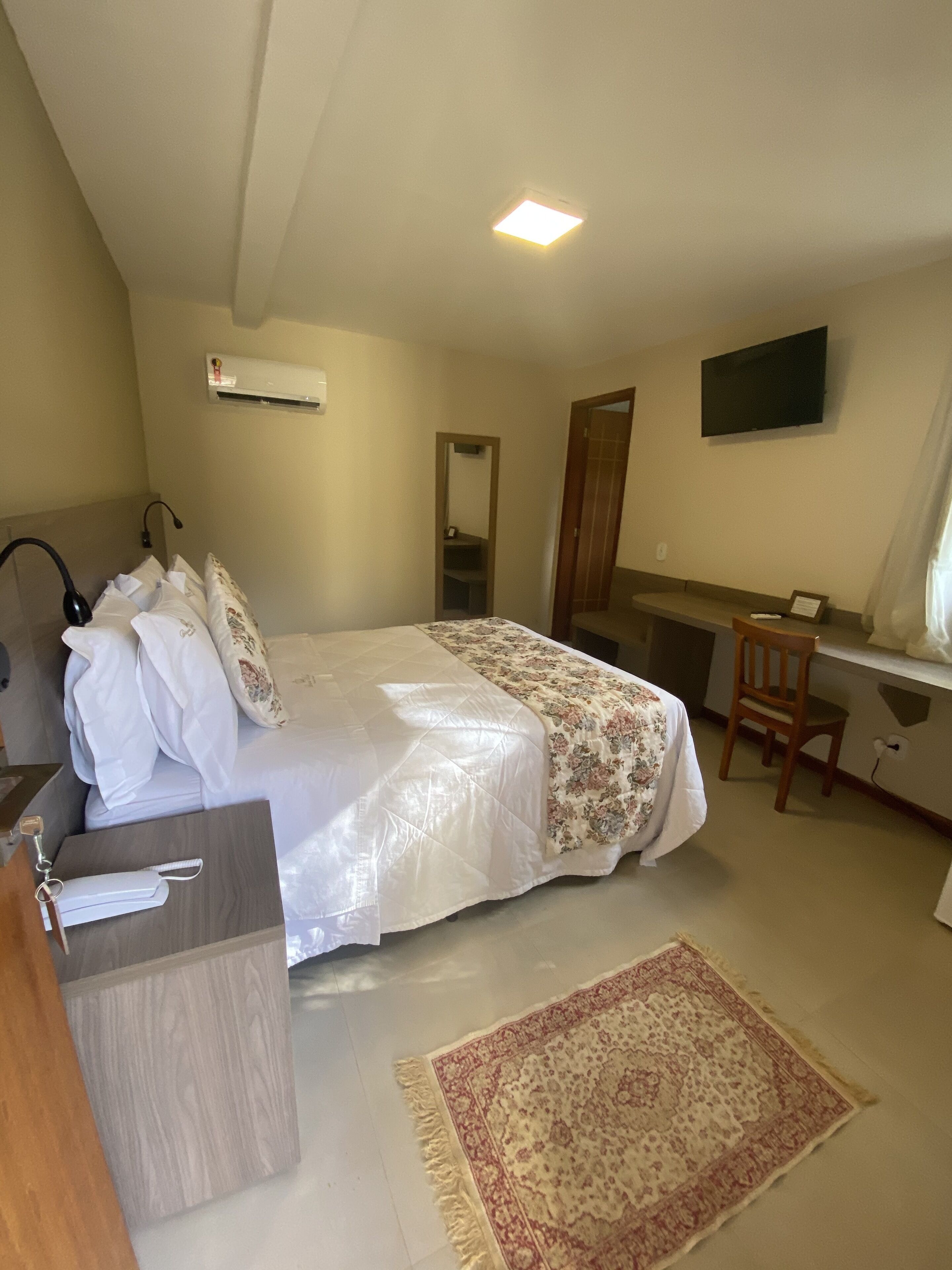 Quarto de hotel com cama, escrivaninha, cadeira, televisão e espelho.