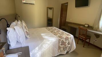Quarto de hotel com cama, escrivaninha, cadeira, televisão e espelho.