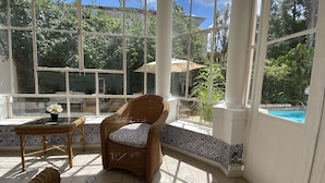 A sunlit room with a wicker chair, a small table with a vase, and a view of a swimming pool through large windows. - Pousada Princesa Isabel - Visconde (Petrópolis)