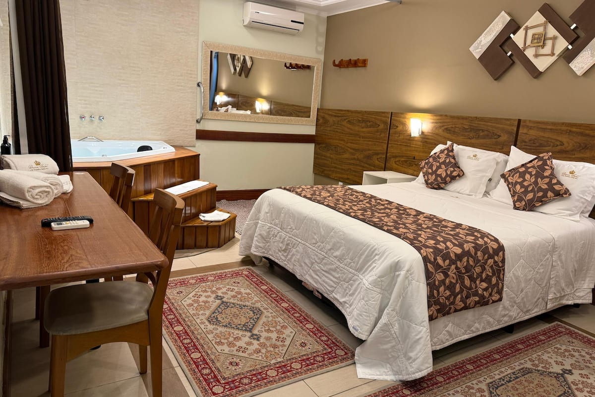 Quarto de hotel com cama, escrivaninha com controle remoto, banheiro com espelho e vista para o exterior.