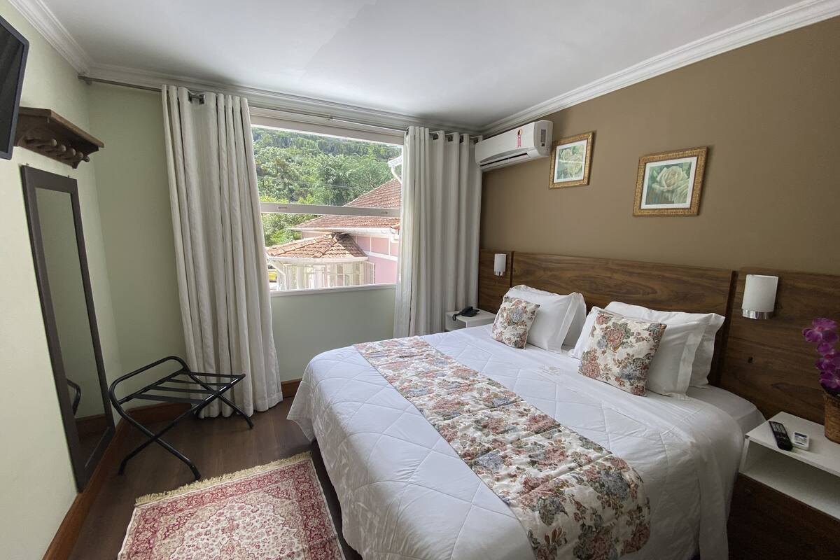 Quarto de hotel com uma cama grande, cabeceira de madeira, mesa de cabeceira com abajur, criado-mudo e uma pequena escrivaninha com telefone, além de uma janela com cortinas.
