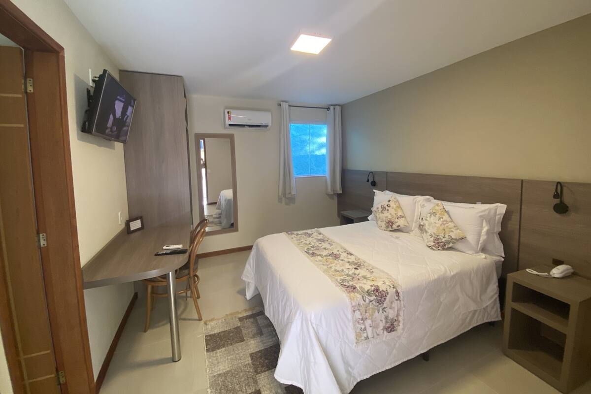 Quarto com cama, mesa de cabeceira, escrivaninha, cadeira, televisão, aparelho de ar condicionado e janela.