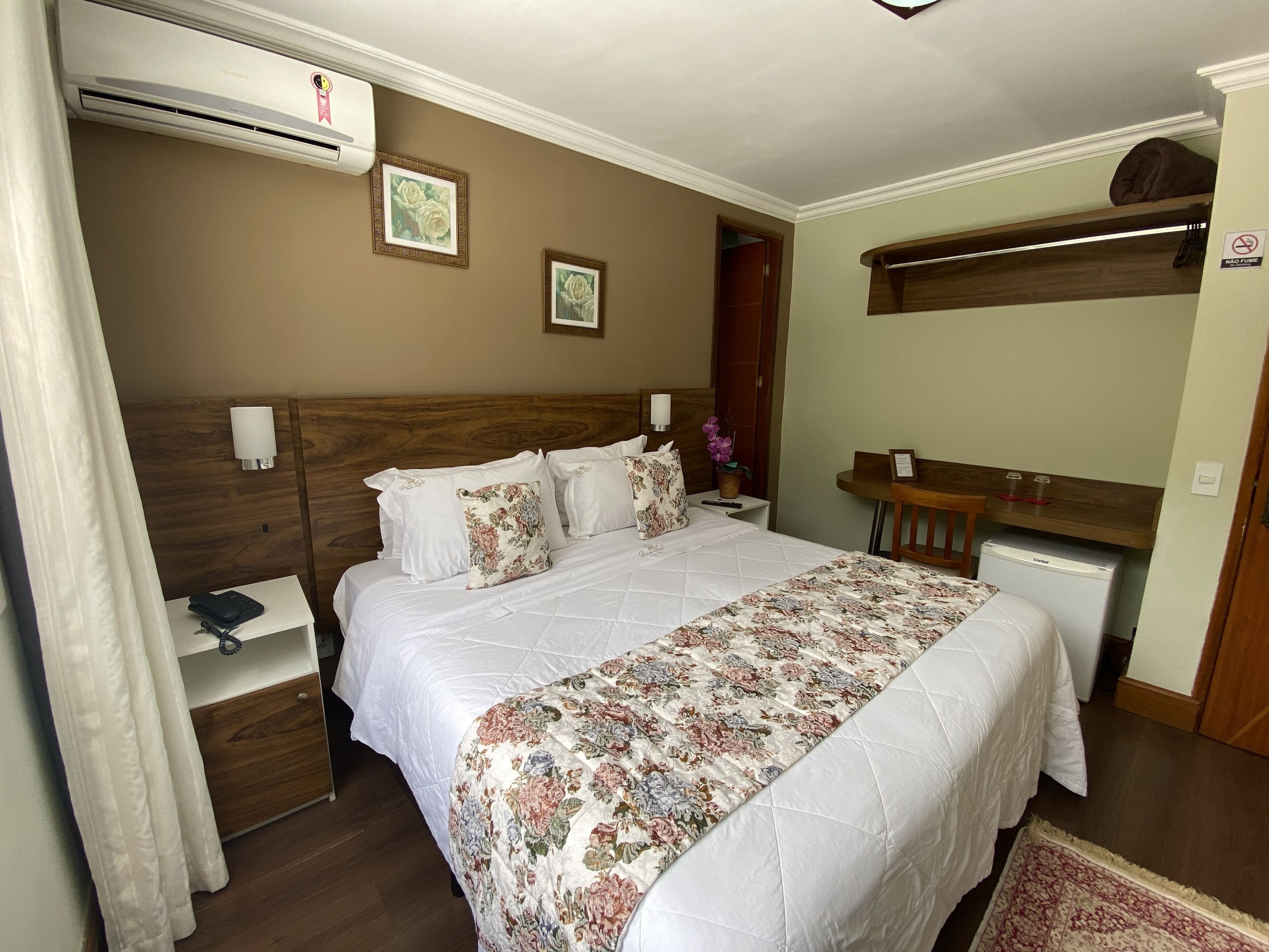 A hotel room with a bed, bedside table, lamp, air conditioning unit, and a small desk with a chair.
