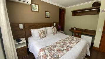 A hotel room with a bed, bedside table, lamp, air conditioning unit, and a small desk with a chair.