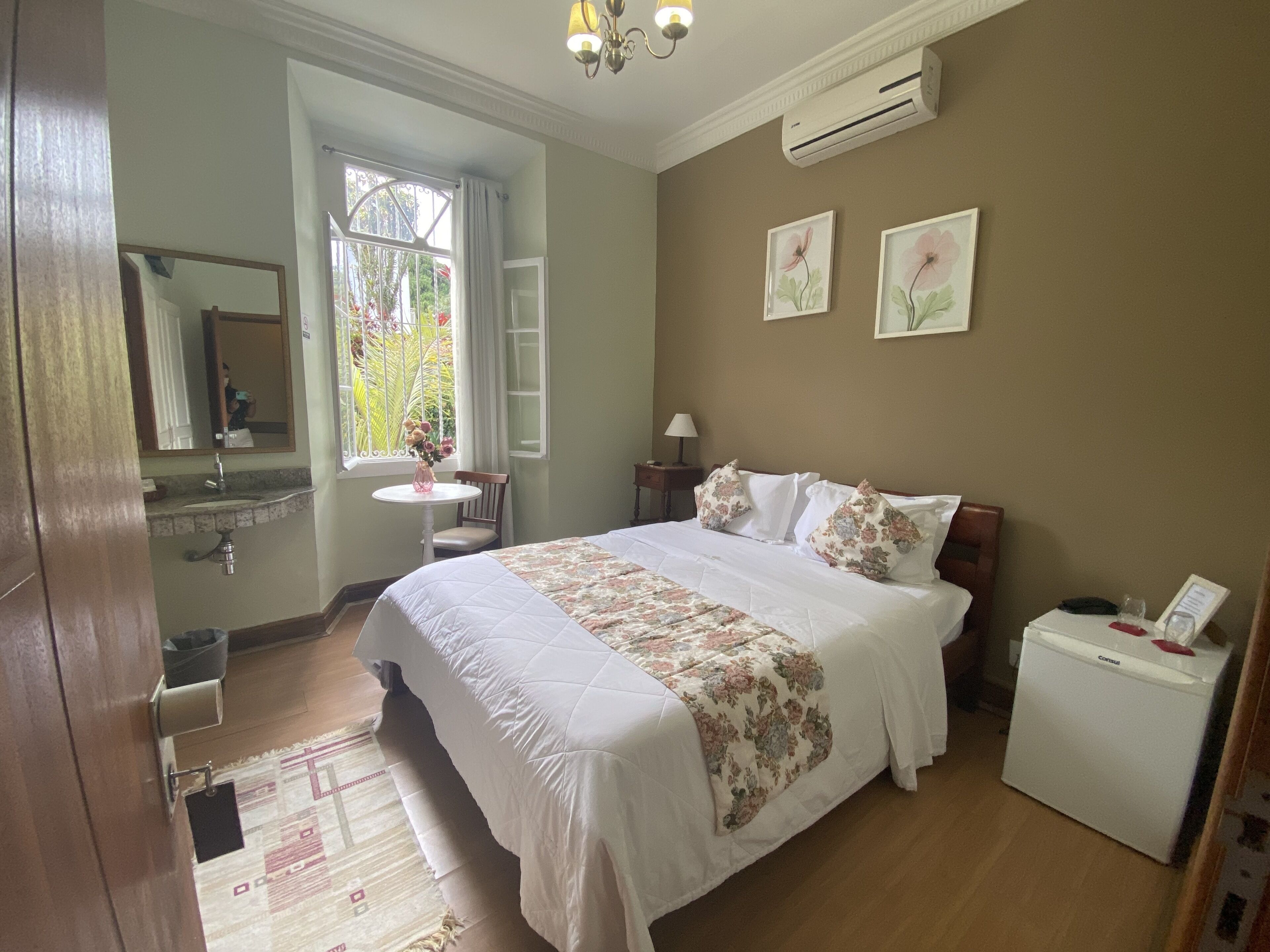 Un dormitorio con cama, mesita de noche, pequeña mesa con lámpara, espejo, aire acondicionado y ventana con cortinas.