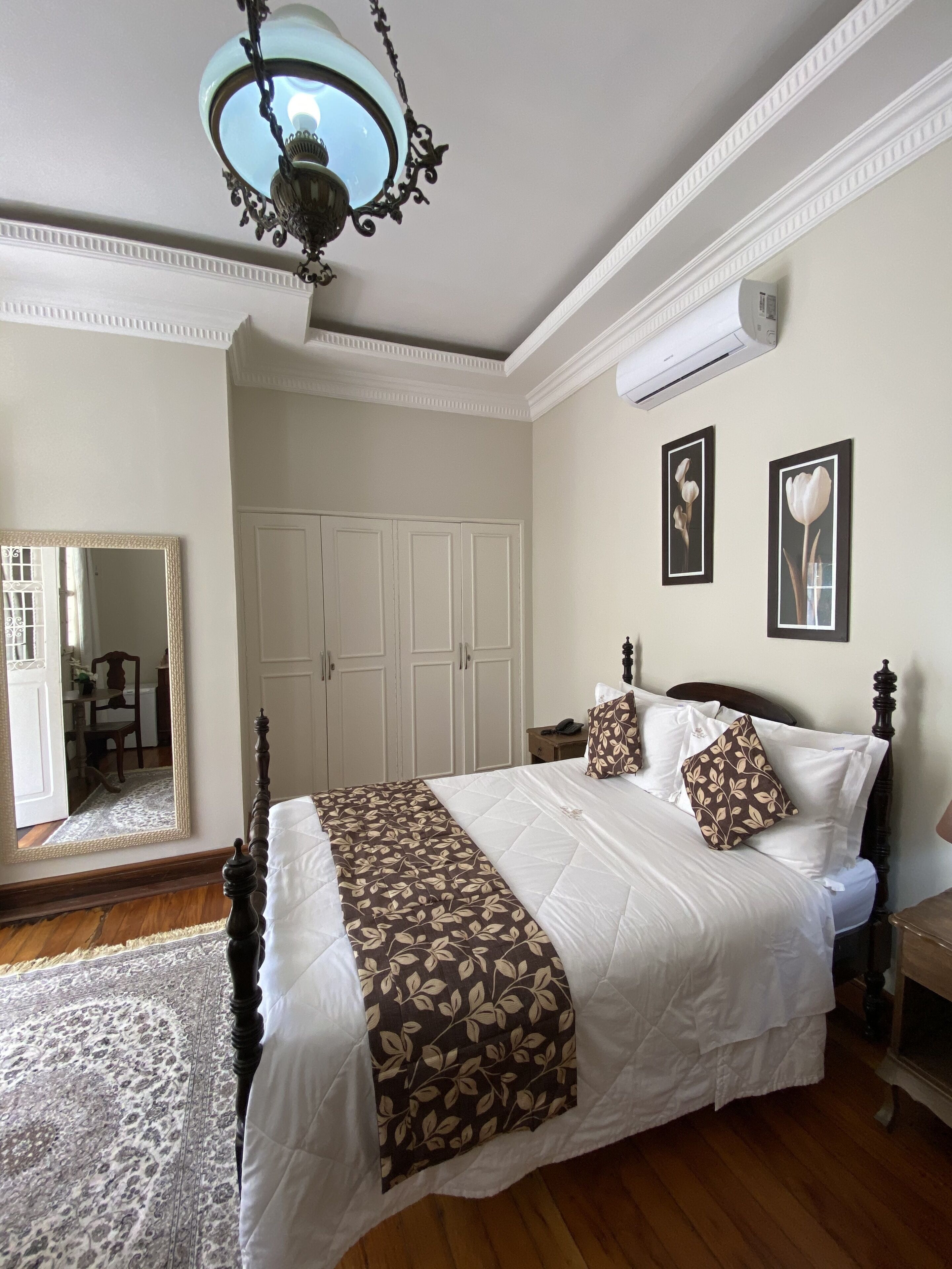 A well-lit bedroom with a large bed, a chandelier, a wardrobe, and two framed pictures on the wall.