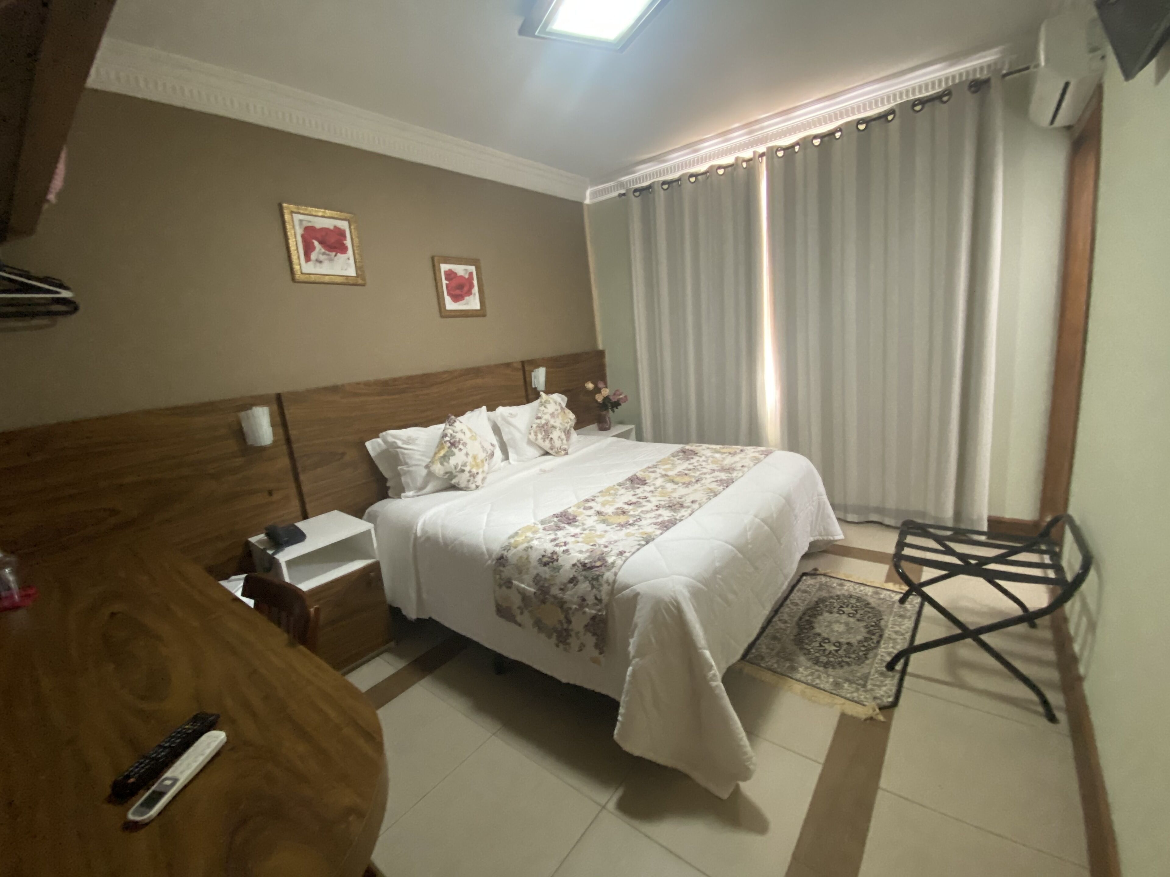 A hotel room with a wooden headboard, a bed with white bedding and a floral throw, a bedside table with a lamp, a small rug, and a window with curtains.