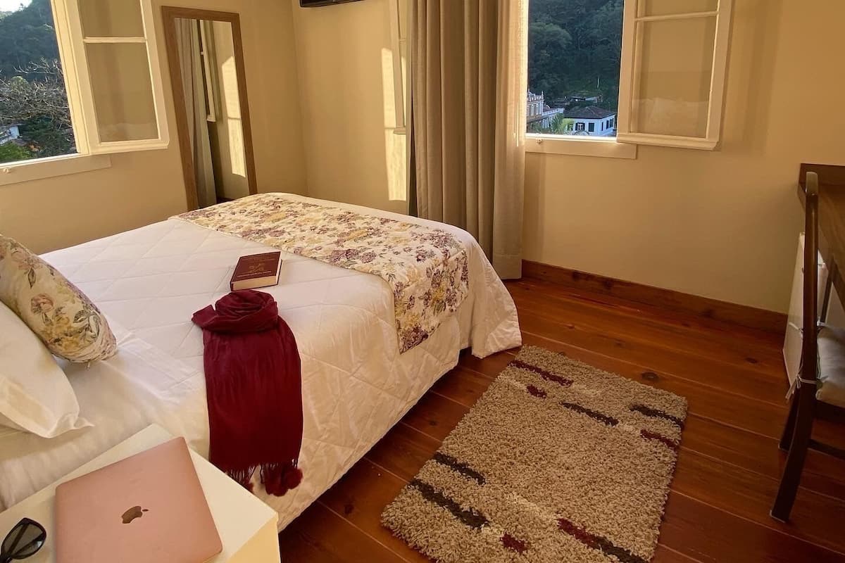 Quarto com cama, laptop, espelho, televisão e vista para uma colina.