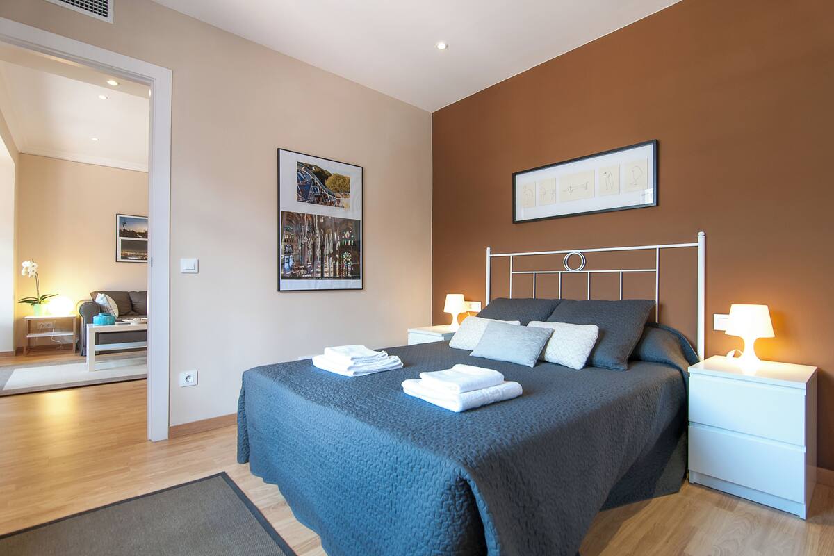A modern bedroom with a large bed, two bedside tables, a nightstand, and a wall-mounted lamp.