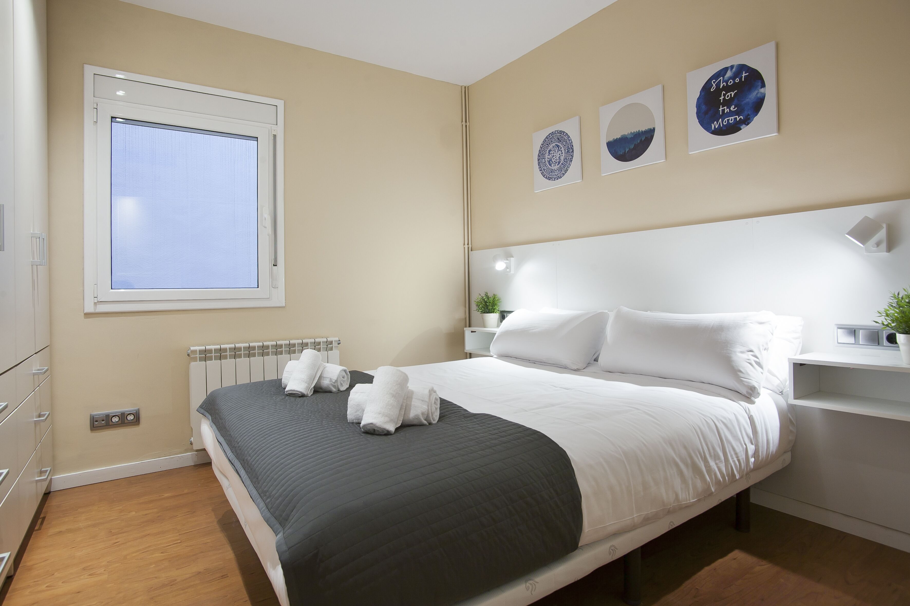 A neatly made bed with white and grey bedding, a window with blinds, and three wall-mounted artworks.