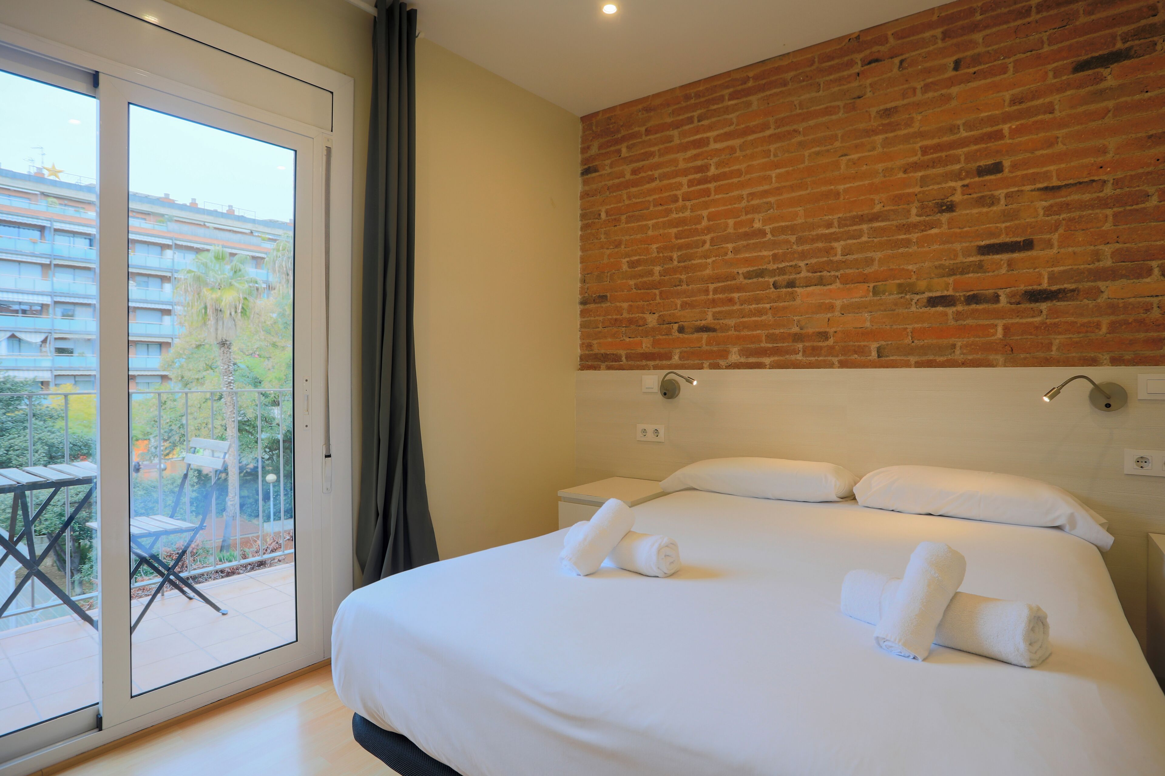 Hotelkamer met een groot bed, twee kussens en twee opgerolde handdoeken. Er is een raam met uitzicht op een balkon en gebouwen.