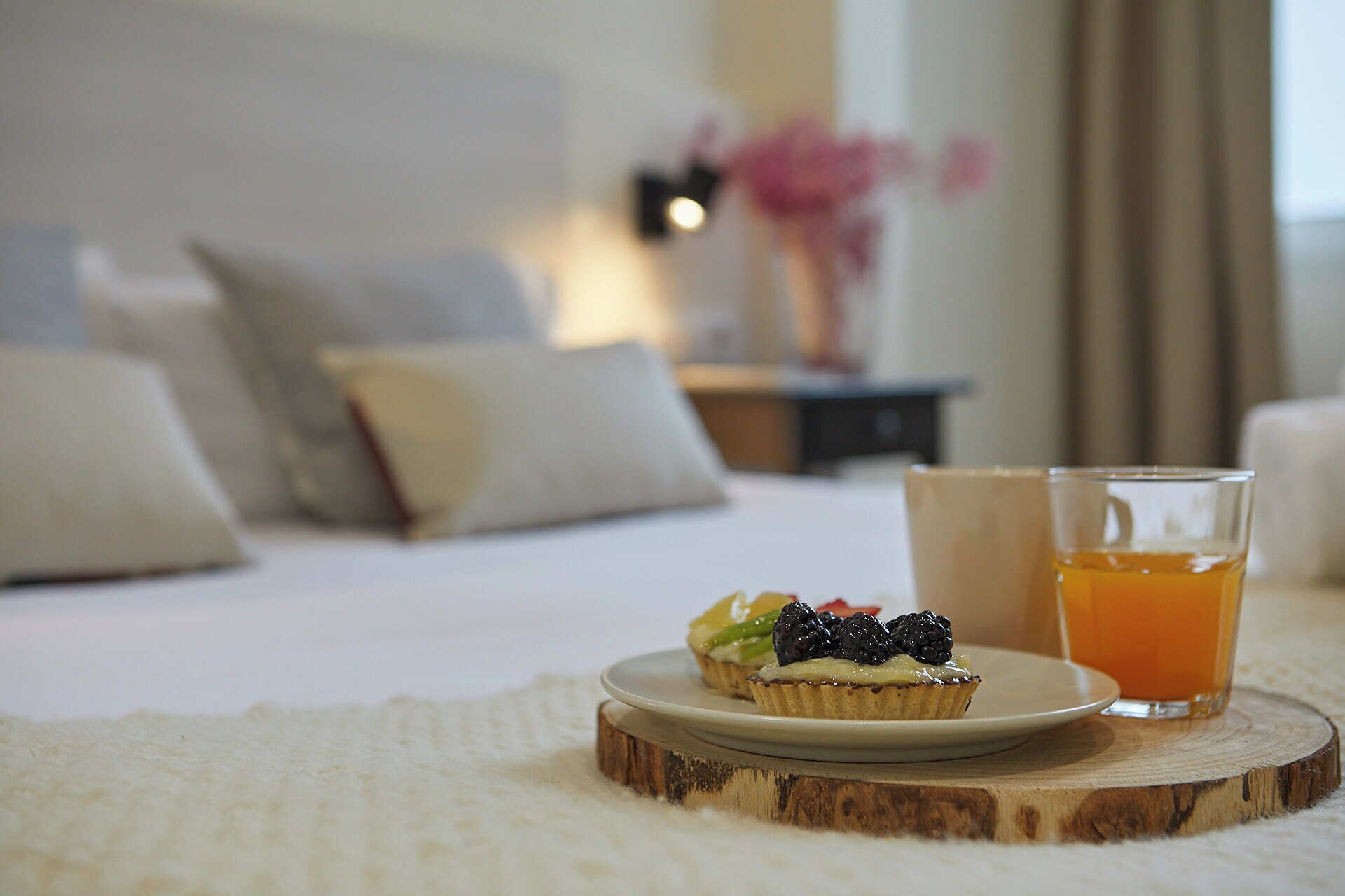 A neatly made bed with pillows, a small plate with a dessert, and a glass of orange juice on a wooden coaster.