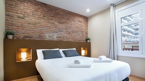 Una habitación de hotel moderna con una pared de ladrillo decorativa, una cama grande con ropa blanca, dos lámparas de noche y vistas a un edificio a través de una ventana.