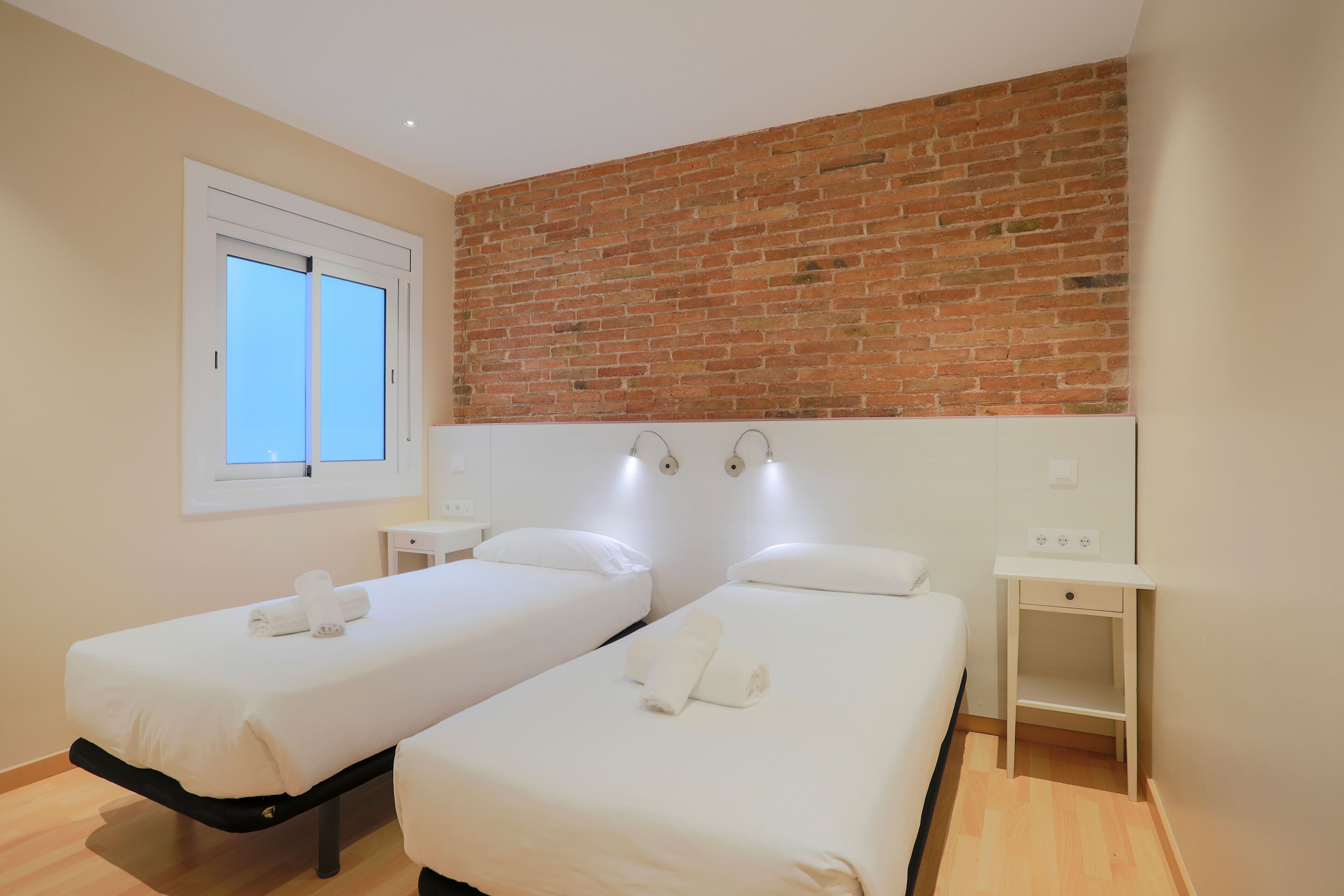 A hotel room with two beds, a brick wall, a window, and a small bedside table.
