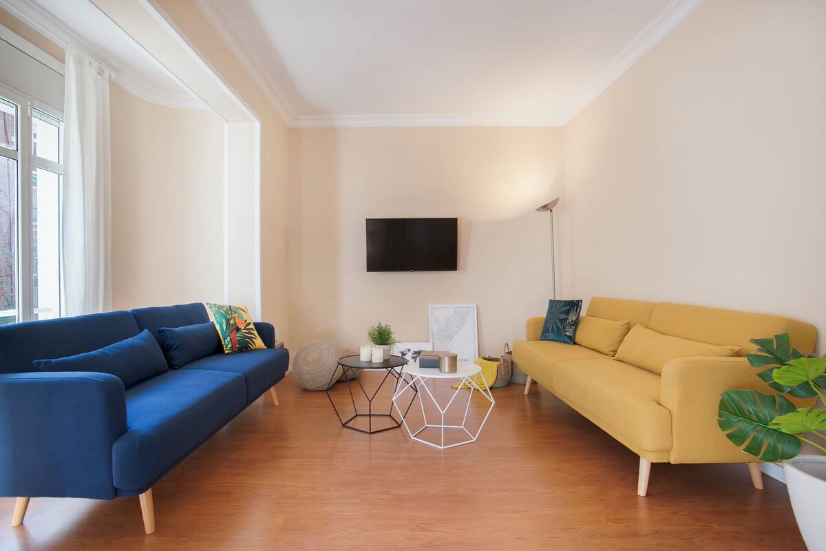A living room with a blue sofa, a yellow sofa, a TV mounted on the wall, and a large window.