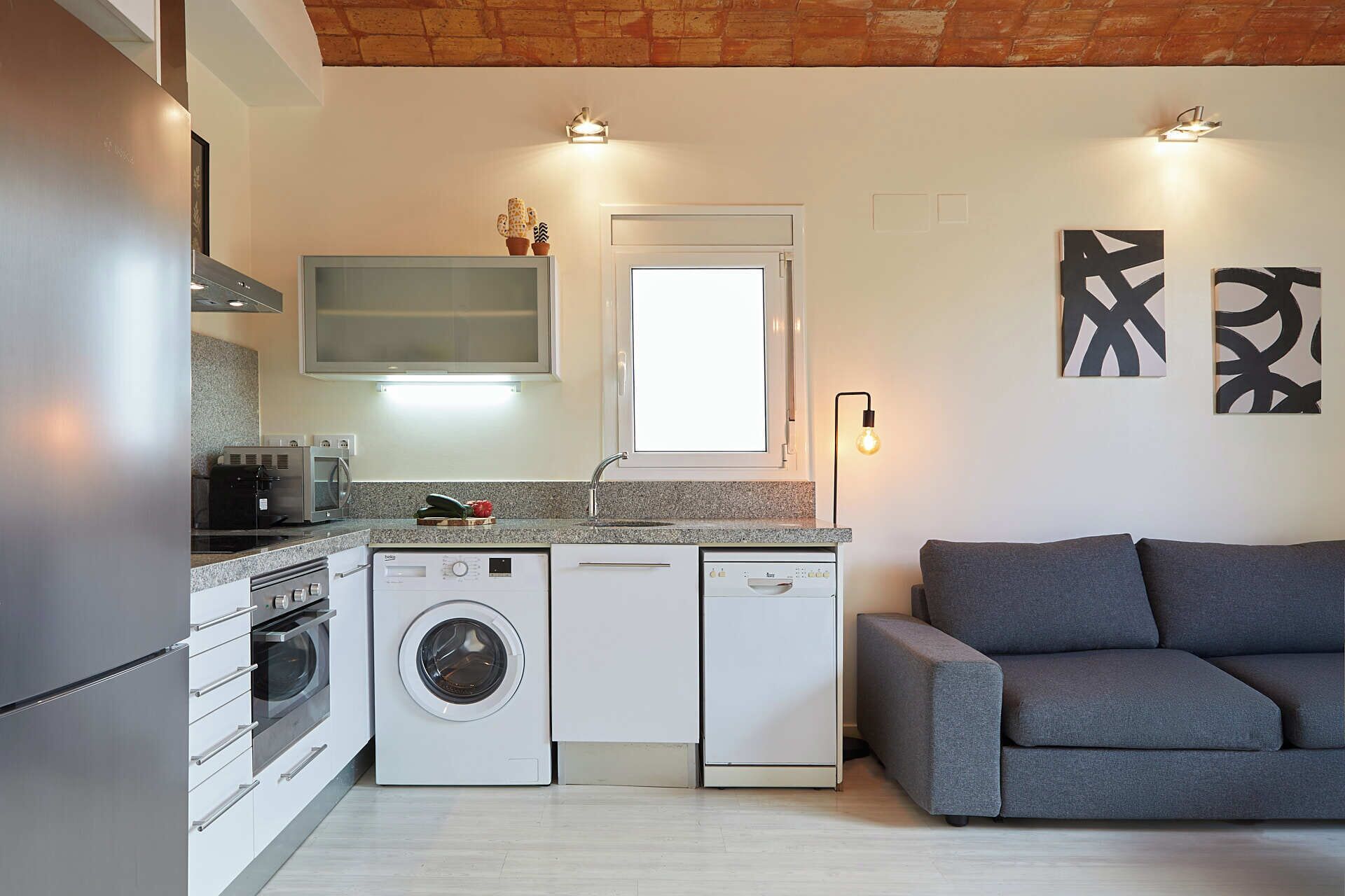Cocina compacta con lavadora, microondas y horno integrado. Hay un sofá gris y una obra de arte abstracta en la pared.