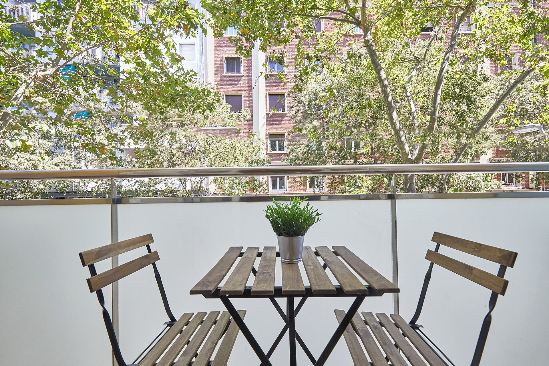 Un balcón con mesa y dos sillas de madera, una planta en maceta sobre la mesa, y vista a un edificio de ladrillo con múltiples ventanas.