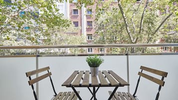 Un balcón con mesa y dos sillas de madera, una planta en maceta sobre la mesa, y vista a un edificio de ladrillo con múltiples ventanas.