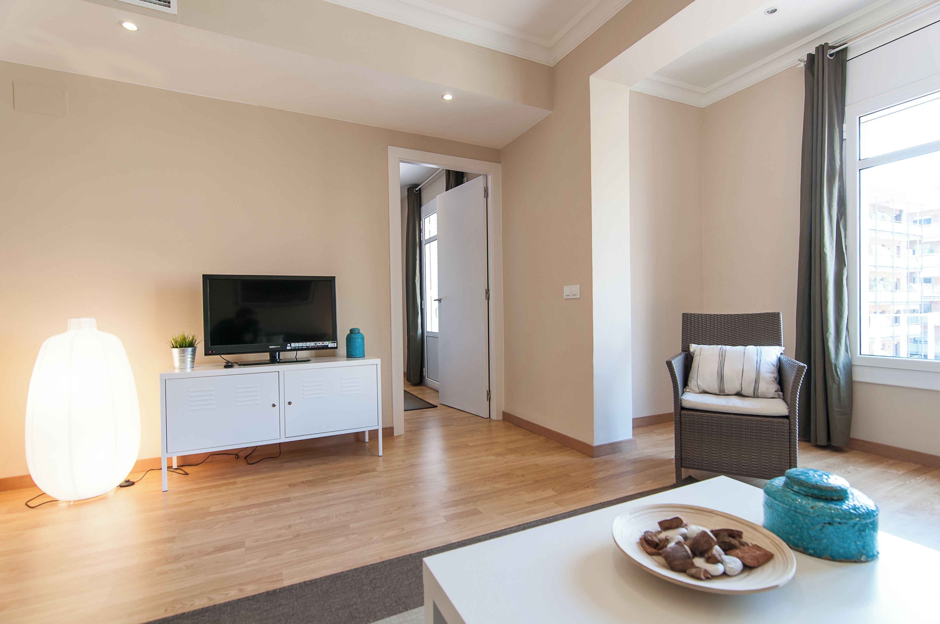 A modern living room with a flat-screen TV, a white cabinet, a wicker chair, a dining table with a plate of food, and a large window with curtains.