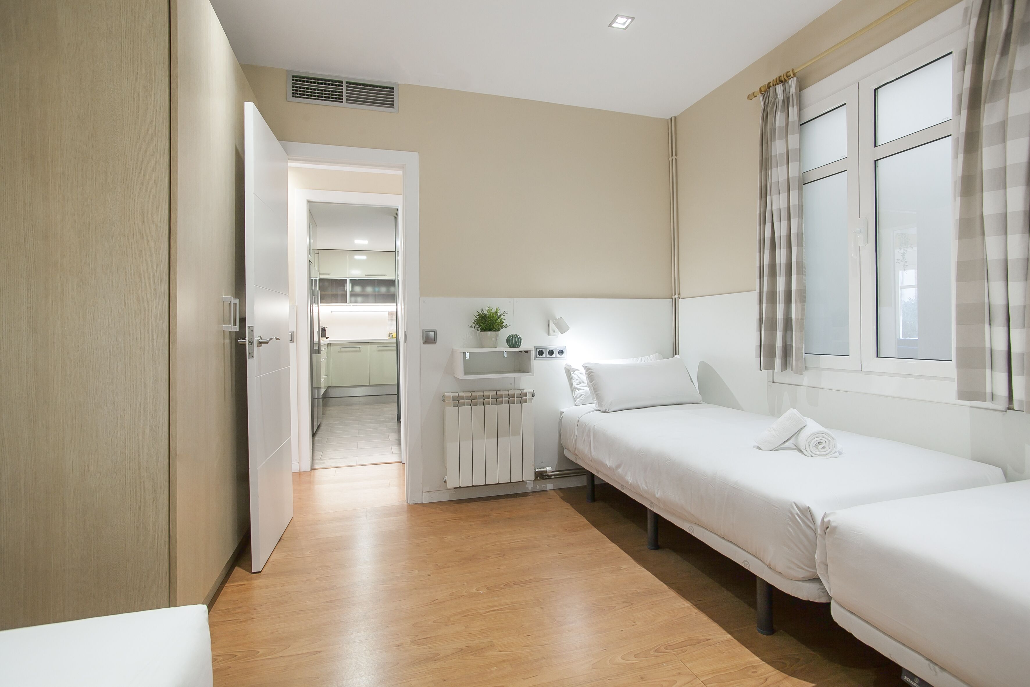 A modern hotel room with a wooden floor, two single beds, a small bedside table with a lamp, a radiator, and a view of the hallway.