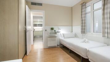 Una habitación de hotel moderna con suelo de madera, dos camas individuales, una mesita de noche pequeña con lámpara, un radiador y vistas al pasillo.