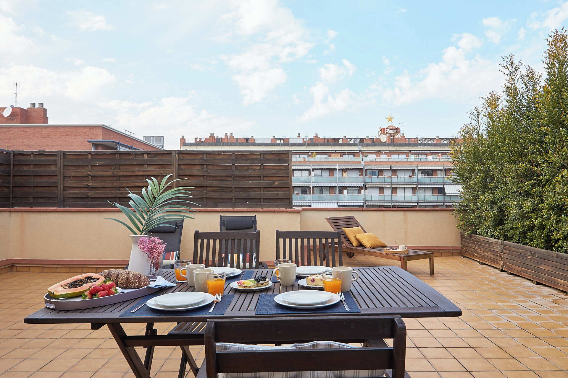 A rooftop terrace with a table set for breakfast, including plates, cutlery, and a variety of fruits and beverages.