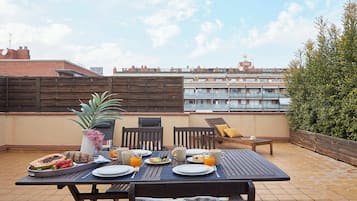 A rooftop terrace with a table set for breakfast, including plates, cutlery, and a variety of fruits and beverages.