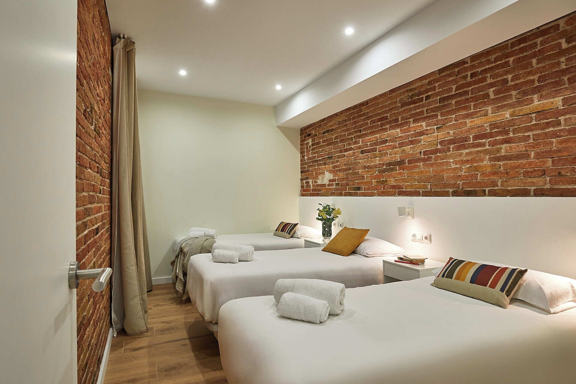 A hotel room with two beds, a brick accent wall, a small bedside table with a vase of flowers, and a curtain on the left.