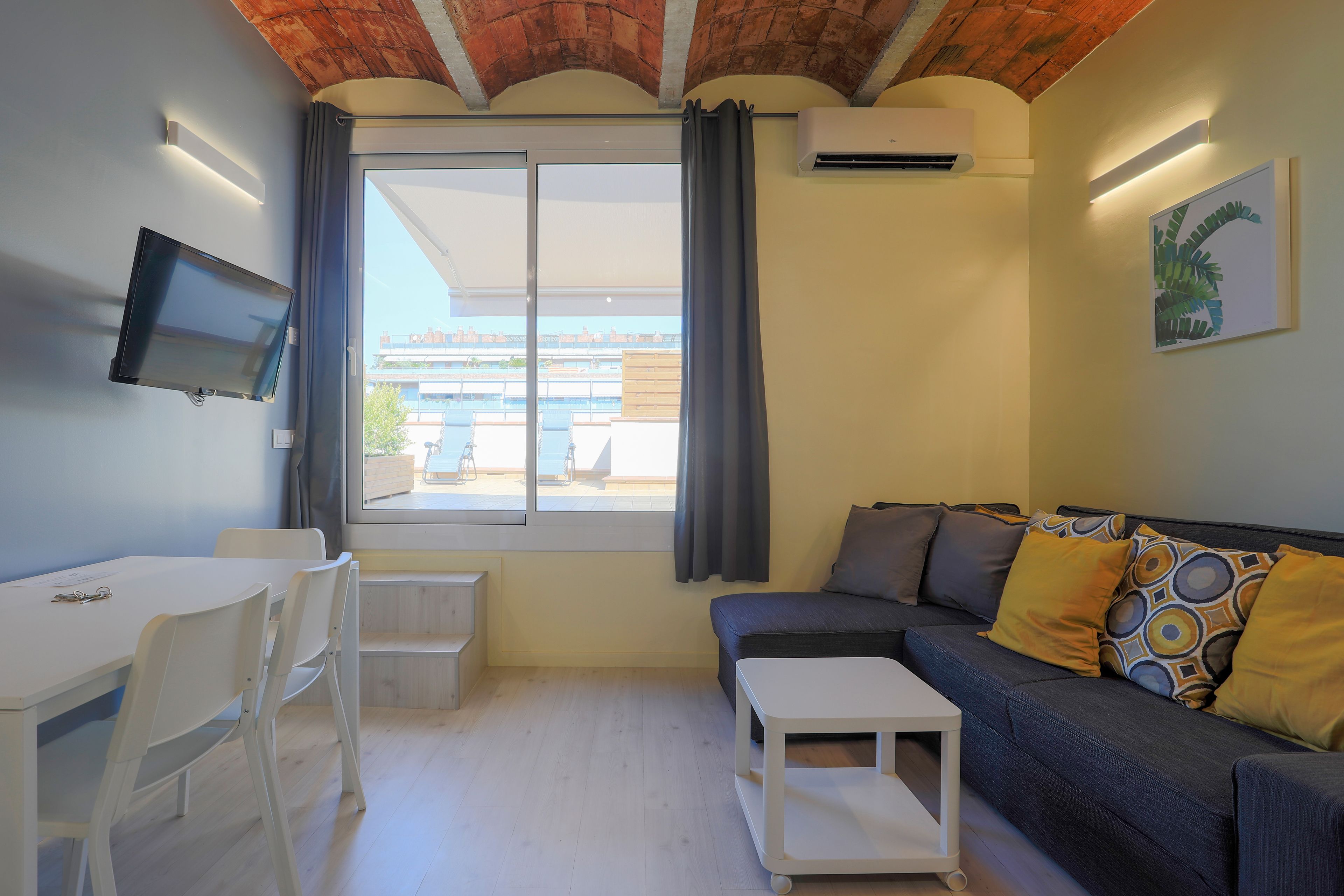 Una sala moderna con sofá, televisor montado en la pared, mesa de comedor con sillas y un ventanal con cortinas.